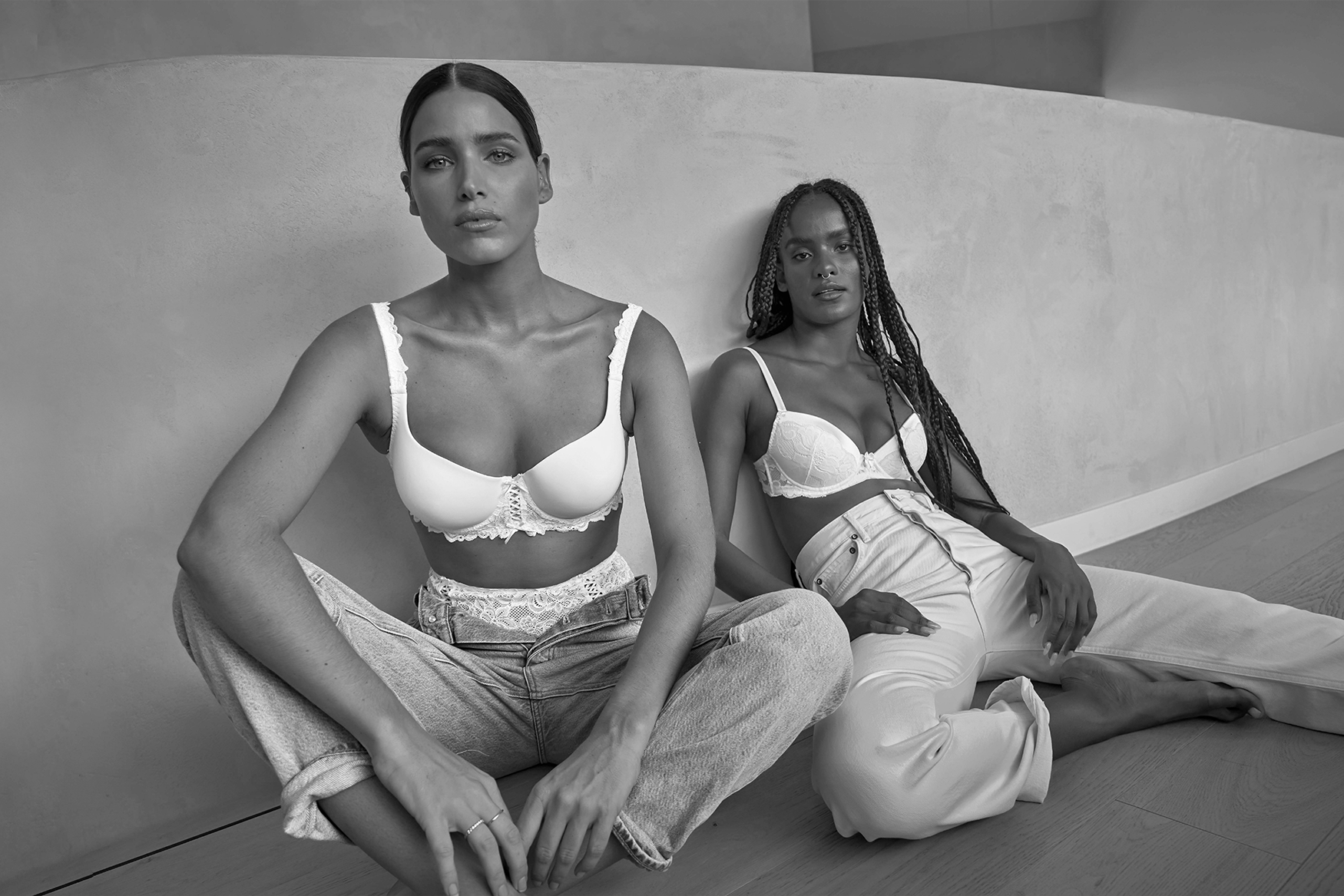 Two women sitting slouching; wearing white lace lingerie and jeans.
